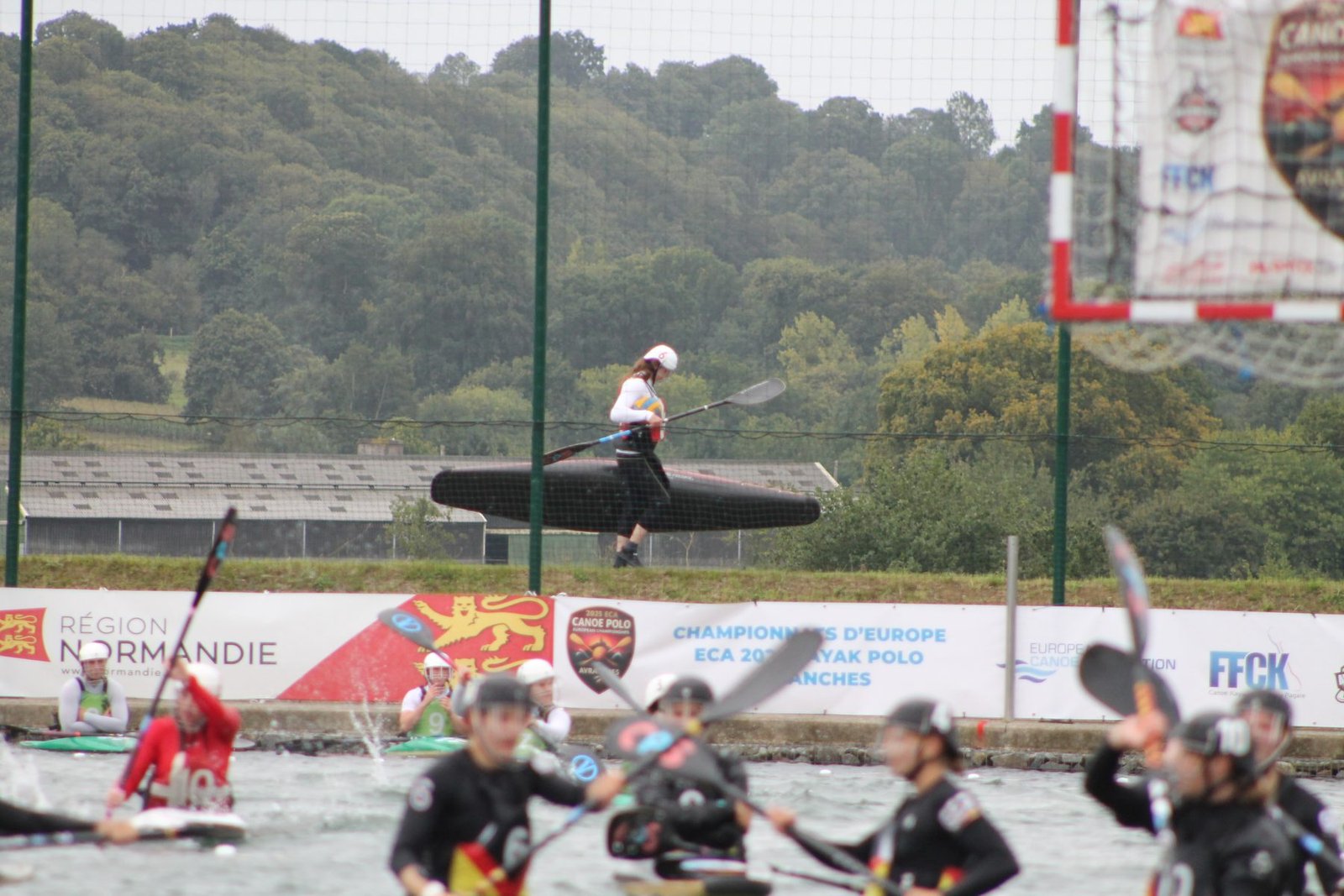 A player carries her kayak past the venue's banners for the ECA 2025 European Championships in Avranches, goal and pitch markings visible.