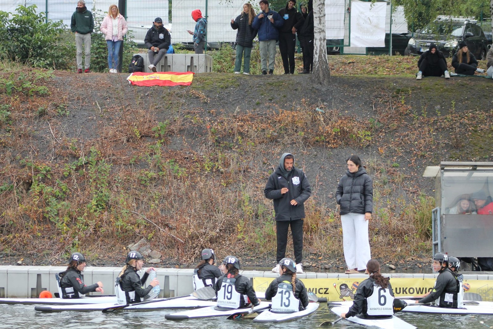 Burriana's kayak polo team sitting in their kayaks by the riverbank in Poland, coaching staff talking on the shore above them, Spanish flag laid on the grass.