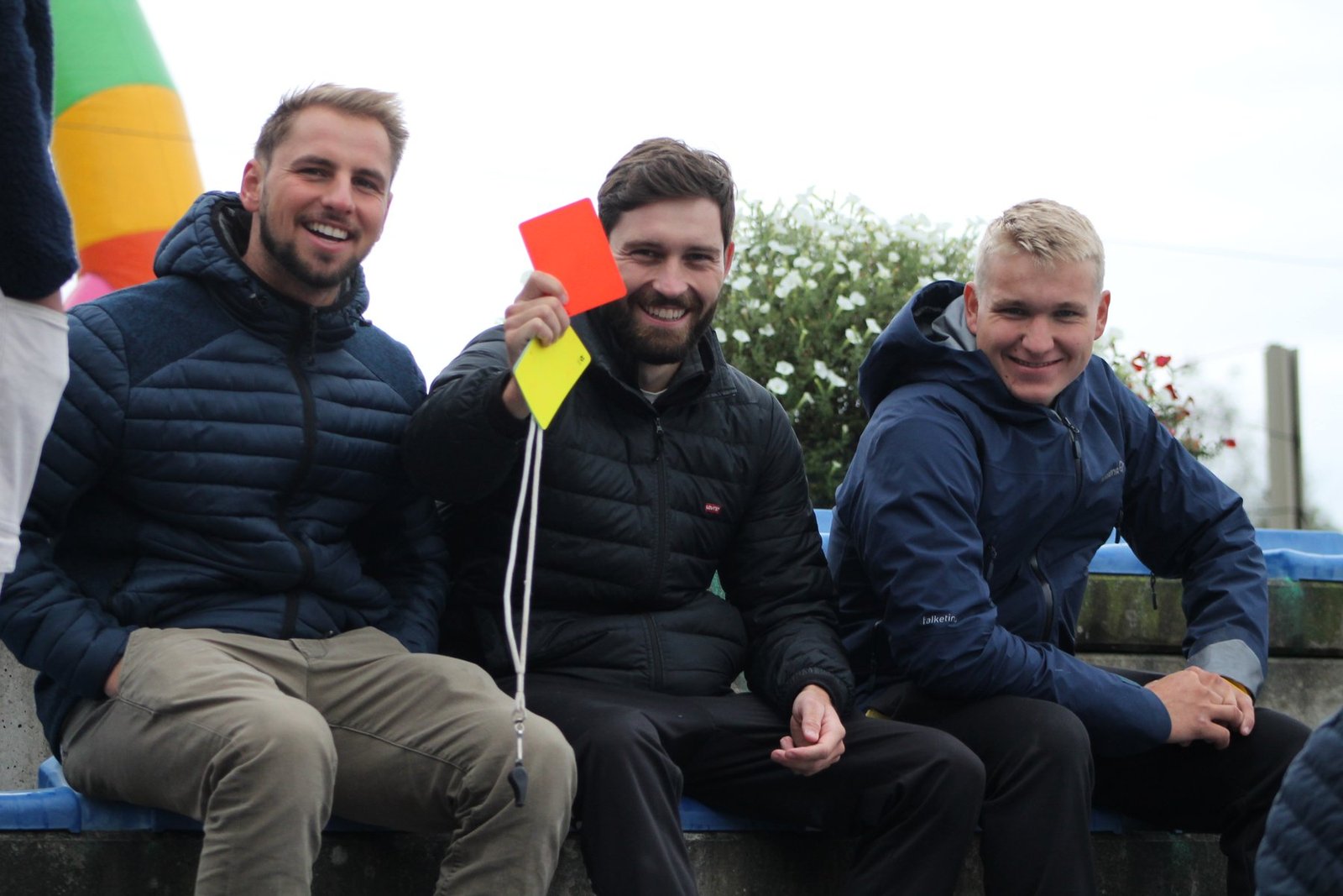 Three smiling players sitting together, one holding up a red and yellow referee card, at an outdoor tournament.