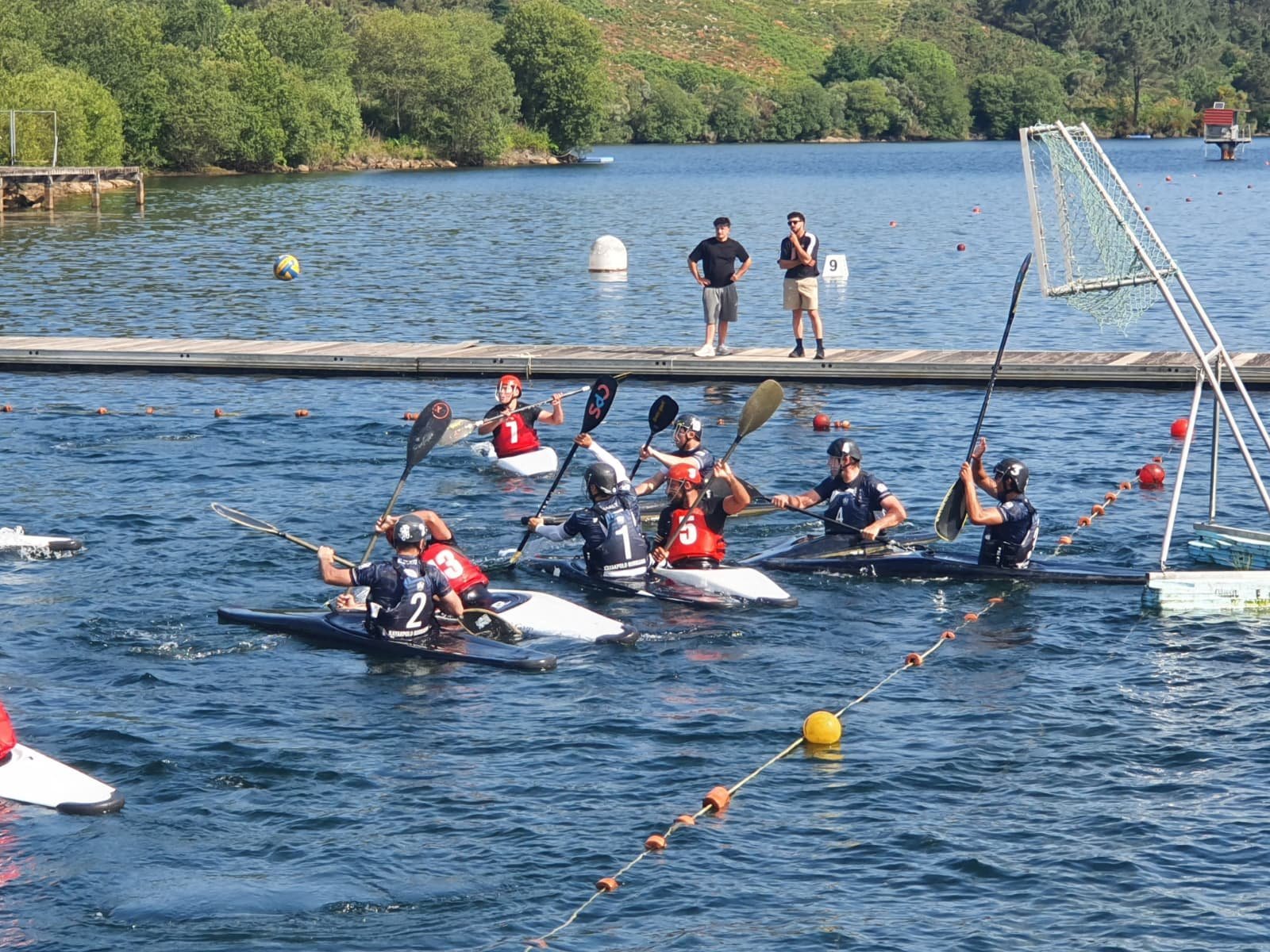 Kayak polo players from Burriana and the opposing team competing for the ball mid-air, near the goal, at the Galicia tournament venue.