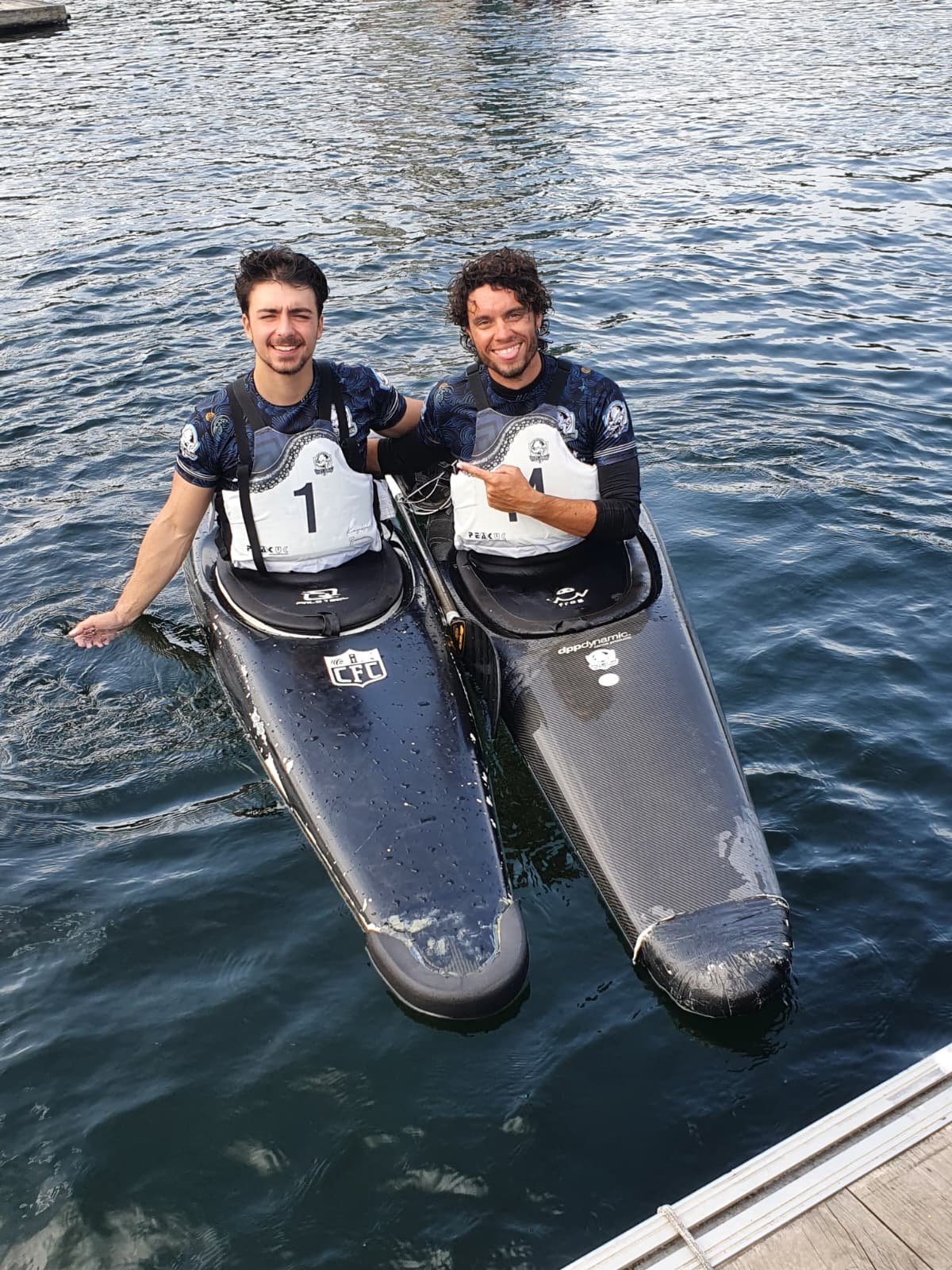 Two kayak polo players resting on their kayaks side by side in the water, smiling, at the Galicia tournament venue.