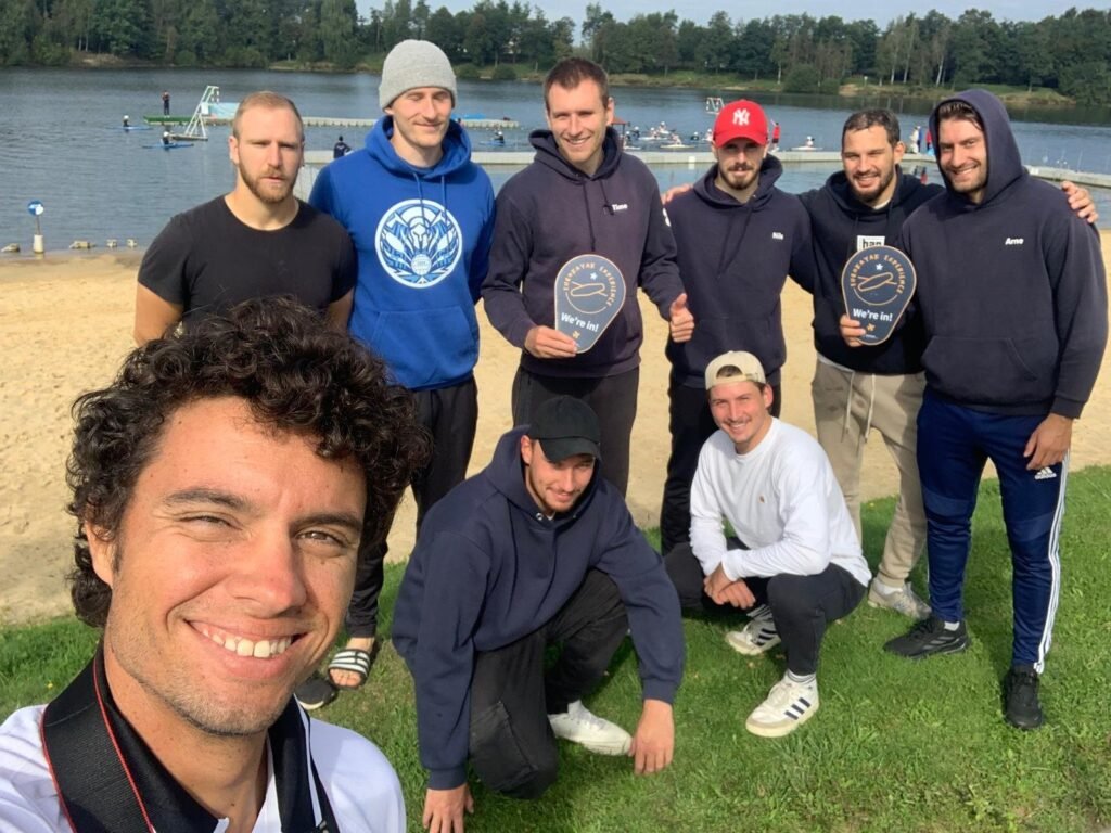 Telmo taking a group selfie with a club's players by a lake, two of them holding 'EuroKayak Experience - We're in!' signs, everyone smiling.