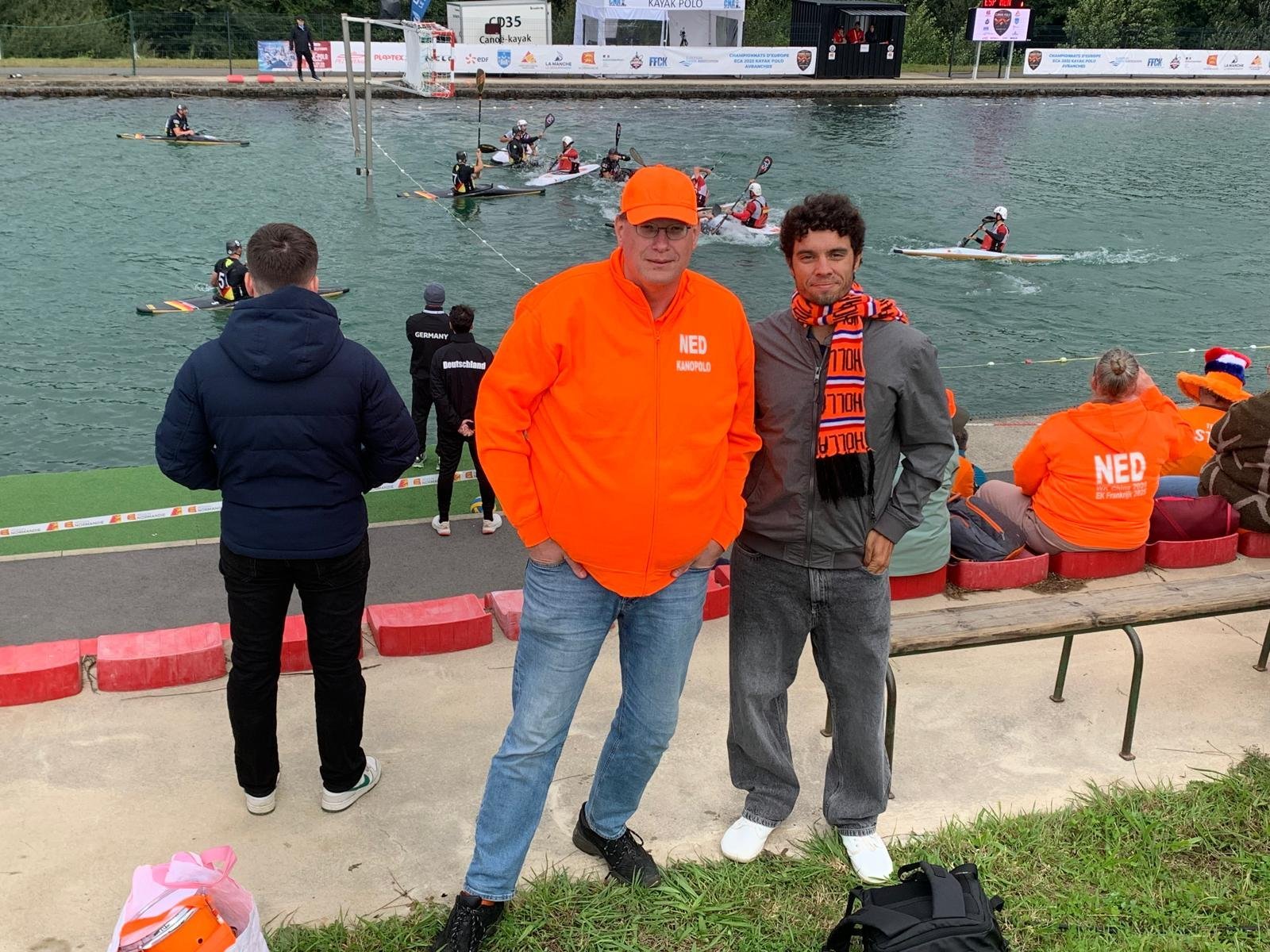 Telmo standing with a Dutch kayak polo coach at the Avranches tournament, both smiling, kayak polo match visible on the water behind them.